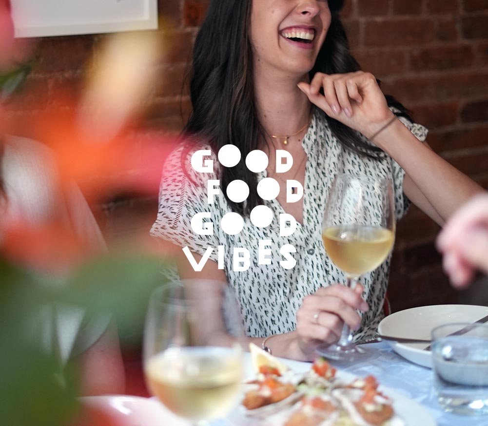 Young woman laughing holding a glass of white wine with oysters on the table with the title GOOD FOOD GOOD VIBES